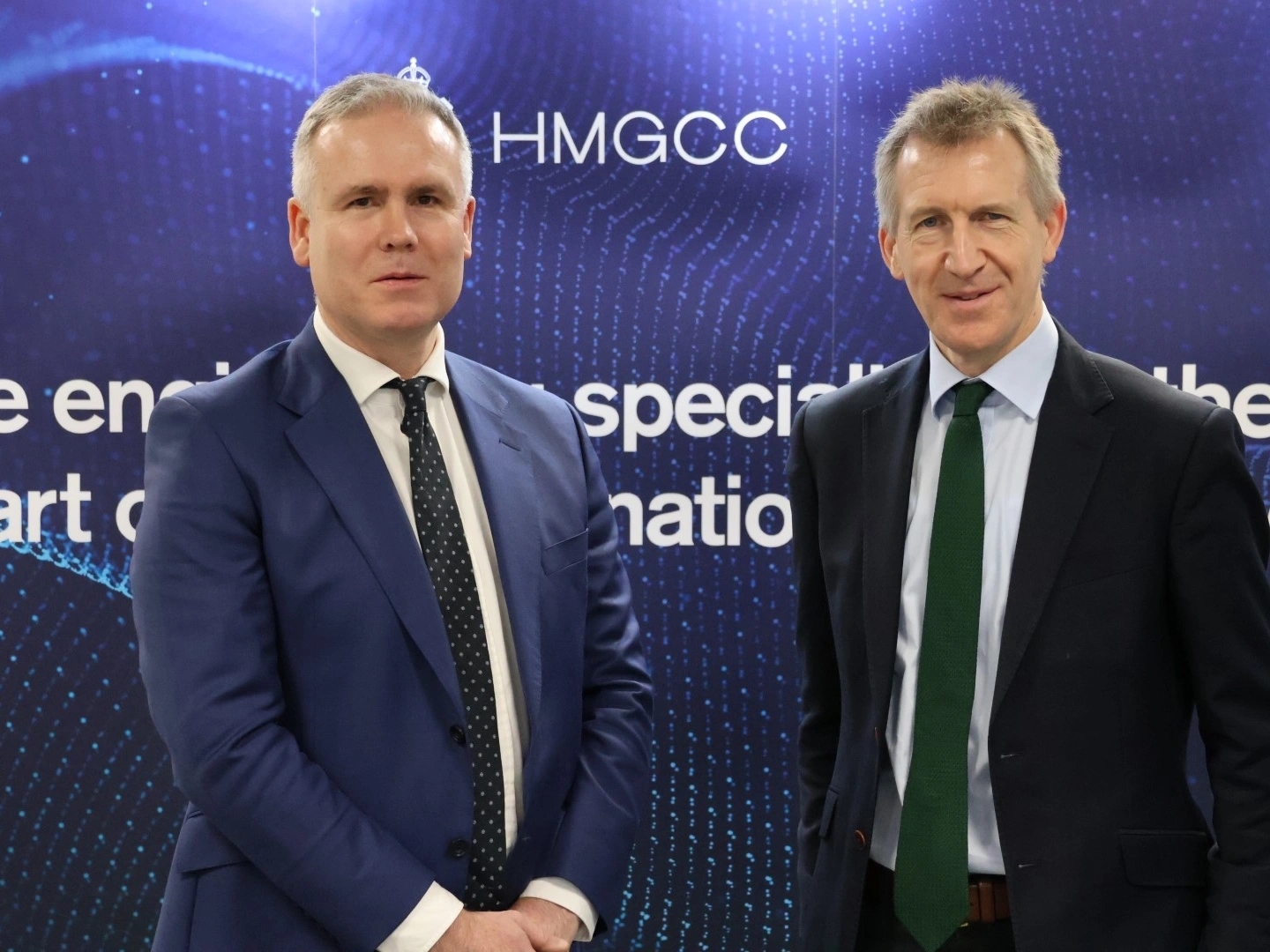 HMGCC CEO George Williamson and Minister of State for Security Dan Jarvis stand in front of an HMGCC poster wall